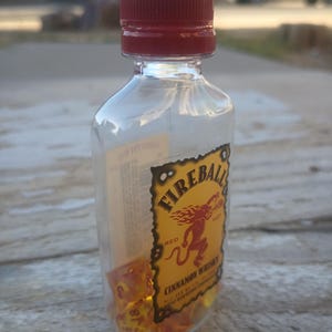 Puede incluir: Una botella de pl&aacute;stico transparente con tapa roja llena de dados naranjas. La botella tiene una etiqueta de Fireball Cinnamon Whisky con una ilustraci&oacute;n de un demonio rojo.