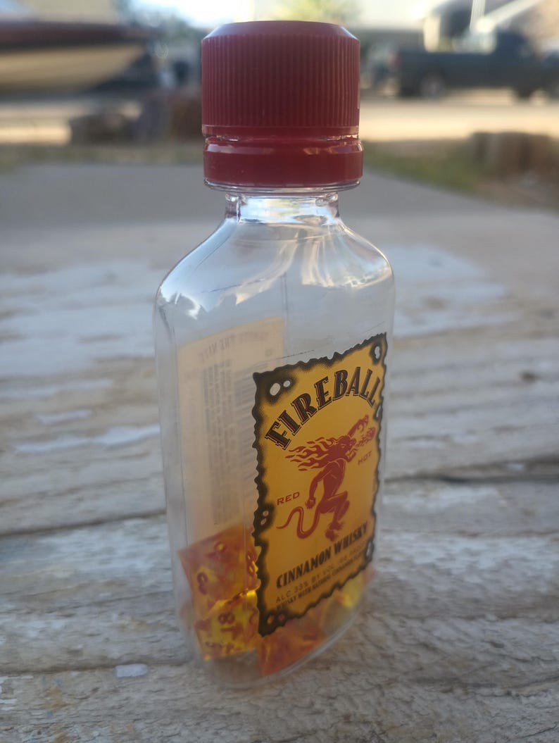 Puede incluir: Una botella de pl&aacute;stico transparente con tapa roja llena de dados naranjas. La botella tiene una etiqueta de Fireball Cinnamon Whisky con una ilustraci&oacute;n de un demonio rojo.