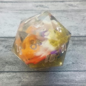 May include: A clear, multi-sided die with embedded orange, purple, and green decorative elements. The die features white numbers and is designed for tabletop gaming. The die is set against a gray wooden surface.