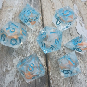 May include: A set of seven clear blue dice with white numbers and copper flecks. The dice are scattered on a light brown wooden surface.