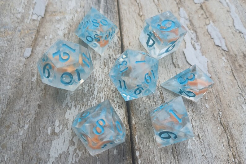 May include: A set of seven clear blue dice with white numbers and copper flecks. The dice are scattered on a light brown wooden surface.
