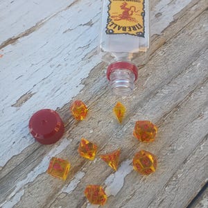 Puede incluir: Un conjunto de dados &aacute;mbar con n&uacute;meros rojos, incluyendo un d4, d6, d8, d10, d%, d12 y d20. Los dados est&aacute;n esparcidos sobre una superficie de madera junto a una botella de vidrio transparente con una tapa roja y una etiqueta que dice "Fireball Cinnamon Whiskey".