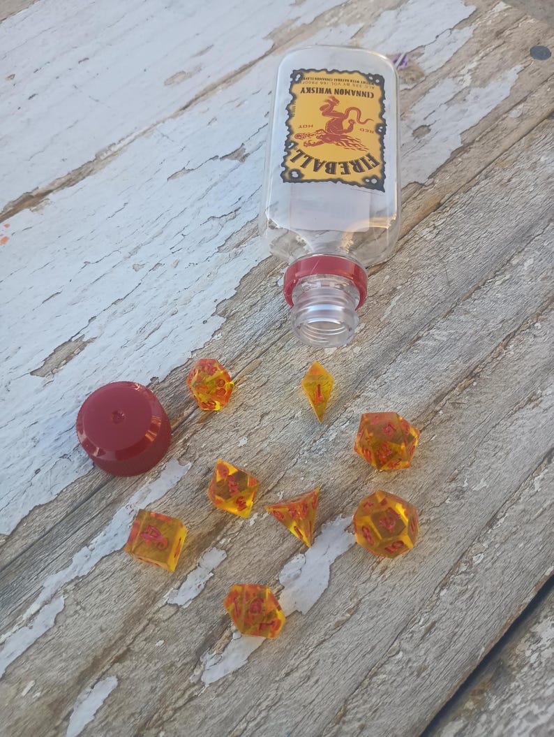 Puede incluir: Un conjunto de dados &aacute;mbar con n&uacute;meros rojos, incluyendo un d4, d6, d8, d10, d%, d12 y d20. Los dados est&aacute;n esparcidos sobre una superficie de madera junto a una botella de vidrio transparente con una tapa roja y una etiqueta que dice "Fireball Cinnamon Whiskey".