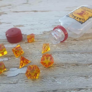 Puede incluir: Un conjunto de dados &aacute;mbar con n&uacute;meros rojos, esparcidos sobre una superficie de madera. Una botella de pl&aacute;stico transparente con una tapa roja y la palabra "Fireball" impresa en la etiqueta est&aacute; tumbada de lado.