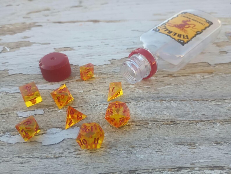 Puede incluir: Un conjunto de dados &aacute;mbar con n&uacute;meros rojos, esparcidos sobre una superficie de madera. Una botella de pl&aacute;stico transparente con una tapa roja y la palabra "Fireball" impresa en la etiqueta est&aacute; tumbada de lado.