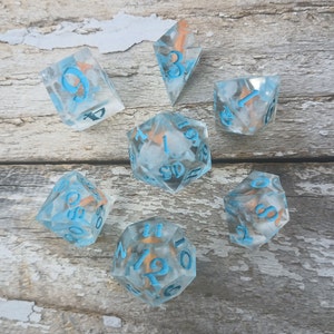 May include: Set of seven clear blue dice with white and gold flecks. The dice are numbered 1 through 6 and have a blue and white swirl pattern. The dice are on a light wood background.