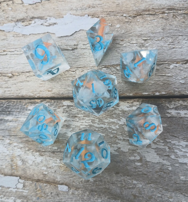 May include: Set of seven clear blue dice with white and gold flecks. The dice are numbered 1 through 6 and have a blue and white swirl pattern. The dice are on a light wood background.