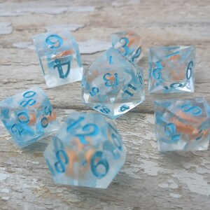 May include: Set of seven blue and white dice with orange flecks. The dice are numbered 1 through 6 and have a variety of shapes, including a d4, d6, d8, d10, d12, and d20.