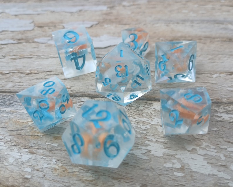 May include: Set of seven blue and white dice with orange flecks. The dice are numbered 1 through 6 and have a variety of shapes, including a d4, d6, d8, d10, d12, and d20.