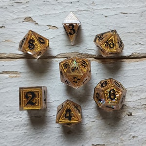May include: Set of seven gold and clear iridescent dice with black numbers. The dice are a d4, d6, d8, d10, d%, d12, and d20.