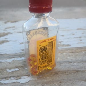 Puede incluir: Una botella de vidrio transparente con una tapa roja llena de dados amarillos. La etiqueta de la botella dice "Ignite the Nite" e incluye el texto "Fireball Whisky" y una direcci&oacute;n de sitio web. La botella est&aacute; sobre una superficie de madera.