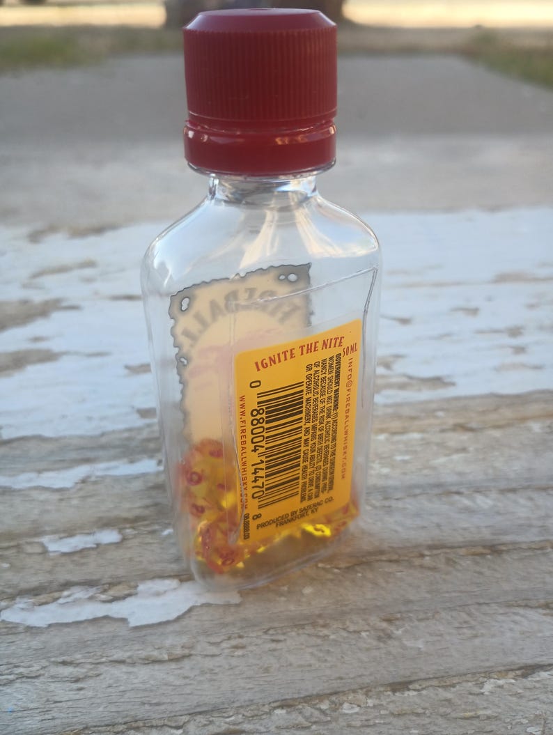 Puede incluir: Una botella de vidrio transparente con una tapa roja llena de dados amarillos. La etiqueta de la botella dice "Ignite the Nite" e incluye el texto "Fireball Whisky" y una direcci&oacute;n de sitio web. La botella est&aacute; sobre una superficie de madera.