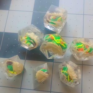 May include: Seven clear, geometric resin dice, each containing a miniature taco. The tacos feature yellow, green, and orange details, encased in transparent resin. The dice are arranged on a checkered surface.