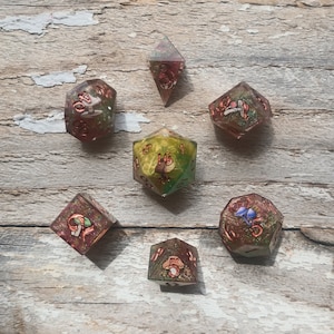 May include: A collection of seven multi-sided dice, each with a unique design and color scheme. The dice are made of clear resin with embedded decorative elements, including copper-colored accents and miniature illustrations. The dice are arranged on a weathered wooden surface.