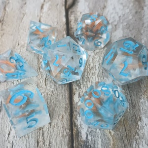 May include: Set of seven clear dice with blue numbers and copper flecks. The dice are on a light wood surface.