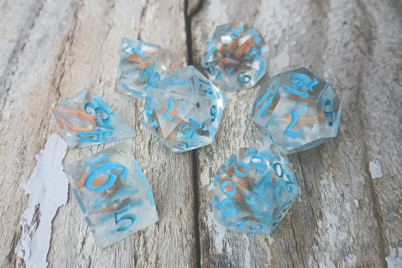 May include: Set of seven clear dice with blue numbers and copper flecks. The dice are on a light wood surface.