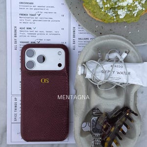 Könnte beinhalten: Eine weinrote Handyhülle mit den Initialen "OS" in goldfarbenen Buchstaben, neben einer Brunch-Speisekarte. Ebenfalls sichtbar sind eine Handcreme-Tube, eine Uhr, eine Haarspange und kabelgebundene Ohrhörer.