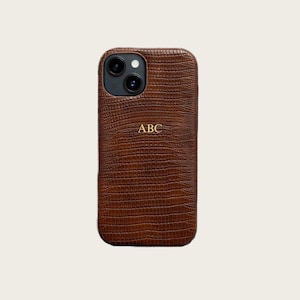 May include: A brown leather phone case with a textured, reptile-like pattern. The case has a gold-colored monogram with the letters "ABC". The phone case has a camera cutout at the top.