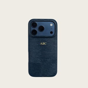 Peut inclure: Une coque de téléphone bleu foncé avec un motif texturé en peau de lézard et une zone de découpe lisse pour l'appareil photo. Le centre de la coque présente les lettres ABC dorées en relief dans une police sans empattement claire et simple.