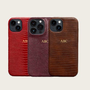 May include: Three personalized phone cases in red, burgundy, and brown. Each case has a textured, reptile-like pattern and gold-colored initials. The cases are designed to fit a smartphone with a multi-lens camera.