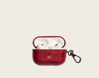 Puede incluir: Un estuche para auriculares inalámbricos de cuero rojo oscuro con textura de cocodrilo, que presenta un monograma dorado con la letra M en el frente. El estuche está abierto, revelando auriculares blancos dentro, con un mosquetón de metal negro.