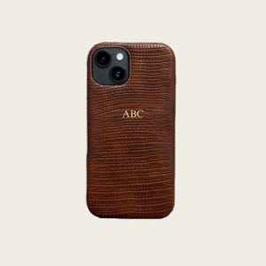 Puede incluir: Una funda de teléfono marrón con un patrón texturizado similar al de un reptil. La funda tiene un monograma dorado con las letras "ABC". La funda del teléfono tiene un recorte para la cámara.