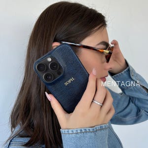 Puede incluir: Funda de teléfono azul marino con un patrón texturizado y el nombre "SOPH." en letras doradas. El teléfono se sostiene cerca de la oreja. La palabra "MENTAGNA" está impresa en el lado derecho de la imagen.