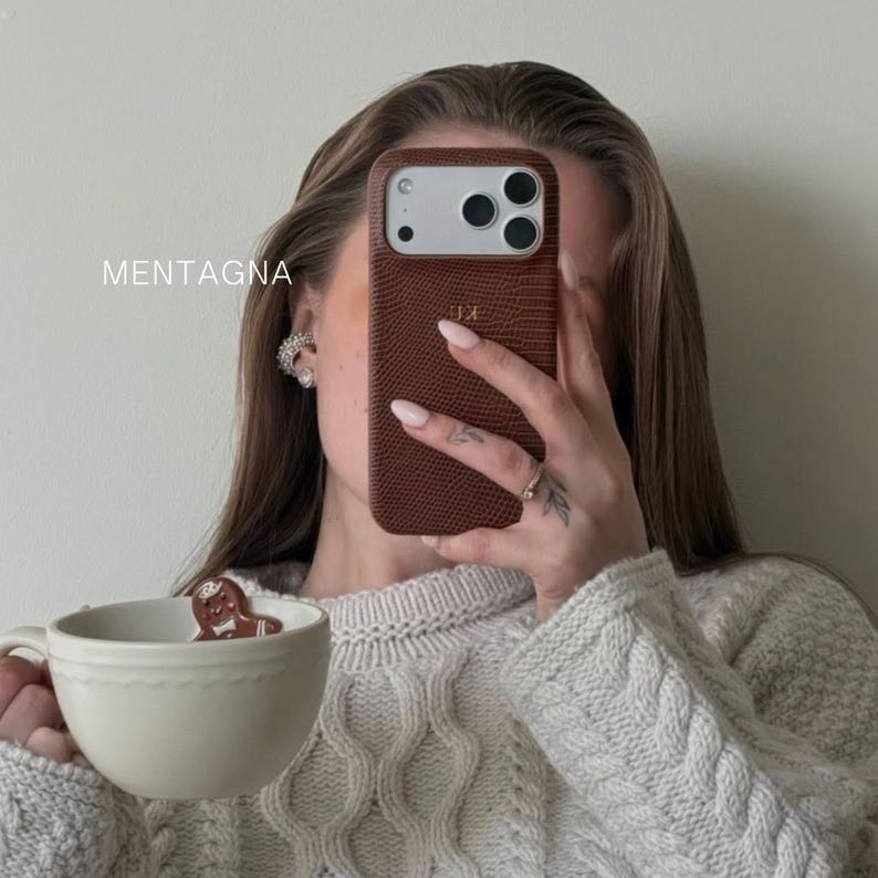 Puede incluir: Una funda de tel&eacute;fono marr&oacute;n texturizada con las iniciales "JK" se sostiene frente a la cara de una persona. La palabra "MENTAGNA" est&aacute; impresa en el lado izquierdo de la imagen. Una taza blanca con un hombre de jengibre se sostiene en la otra mano.