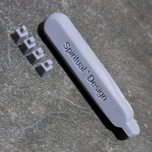 May include: A light gray tool with the text "Spiritual Design" printed on it, along with three small, matching gray square pieces. The tool has a long, rectangular shape with a rounded end.