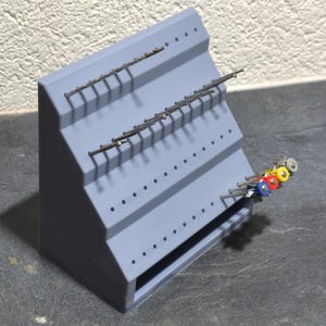 Peut inclure: Un présentoir gris à plusieurs niveaux avec plusieurs rangées de petits trous. Plusieurs broches métalliques sont insérées dans les trous, tenant divers petits objets. Quelques objets avec des dessus colorés sont visibles.