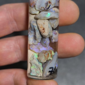 Könnte beinhalten: Ein kleines Glasfläschchen mit goldenem Deckel, gefüllt mit rohen Opalsteinen. Die Opale haben eine Mischung aus Farben, darunter Weiß, Blau und grüne Blitze. Auf dem Fläschchen stehen schwarze Zahlen.