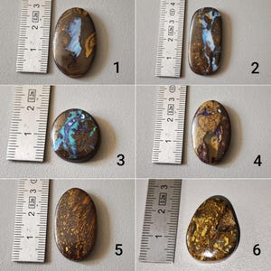 Peut inclure: Une collection de six pierres d'opale polies, chacune avec des motifs et des couleurs uniques. Les opales varient en forme, d'ovale à rectangulaire, avec une règle à côté de chaque pierre pour la référence de taille. Les opales affichent une variété de couleurs, dont le bleu, le marron, l'or et le vert.