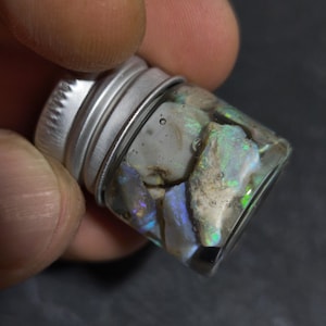 Peut inclure: Un petit flacon en verre transparent avec un bouchon argenté, rempli de fragments d'opale irisés. Les opales affichent des éclats de bleu, de vert et de violet. Le flacon est tenu dans une main sur un fond sombre.