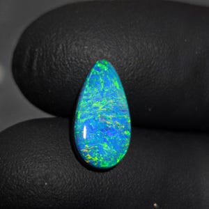 Könnte beinhalten: Ein tropfenförmiger Opal-Edelstein mit einem lebendigen Farbspiel. Der Stein hat eine tiefblaue Basis mit grünen und gelben Lichtreflexen. Die polierte Oberfläche reflektiert das Licht und zeigt die inneren Muster und Texturen des Opals.