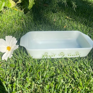 Puede incluir: Una fuente rectangular Pyrex verde claro con un diseño floral, sobre hierba verde. La fuente tiene una forma ligeramente curvada y asas en cada extremo. Flores blancas con centros amarillos están dispersas alrededor de la fuente.
