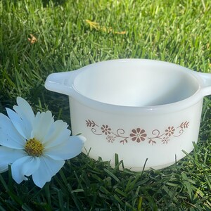 Puede incluir: Una fuente para horno Pyrex blanca y redonda con dos asas y un diseño floral marrón. Una flor blanca con centro amarillo está junto a la fuente. La fuente está sobre hierba verde.