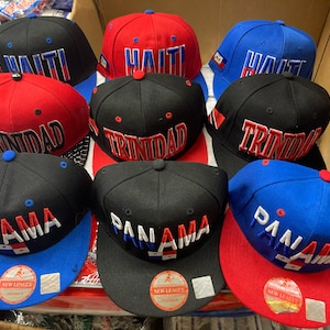 May include: Nine baseball caps with different country names and flags embroidered on them. The caps are black, red, and blue with white lettering. The caps are arranged in three rows of three. The caps are all new and have tags attached.