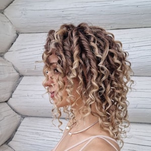 May include: A woman with long, curly, light brown hair with blonde highlights. She is wearing a light pink, sleeveless top with a thin silver necklace.