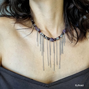 Peut inclure: Un collier avec un design de perles avec des pierres violettes et rouges, entrecoupées de perles argentées. Des franges de chaîne argentées pendent de la section perlée. Le collier est porté sur un haut sombre.