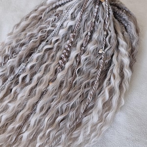 Puede incluir: Una colección de rastas grises y blancas con una textura ondulada. Algunas rastas están trenzadas y otras adornadas con pequeñas cuentas y abalorios. Las rastas están atadas en la parte superior con una pequeña etiqueta.