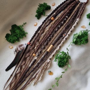May include: A selection of brown and tan dreadlocks, some with decorative beads and woven details, attached to a brown hair tie. A feather and sprigs of green moss and leaves are arranged around the dreadlocks.