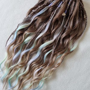 May include: Brown and blonde synthetic dreadlocks with streaks of blue, white, and green. The dreadlocks are styled with wavy ends and some are braided. A small heart charm and other decorative elements are attached.