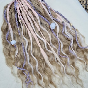 Puede incluir: Un conjunto de extensiones de cabello rubio ondulado con mechones trenzados en rosa claro y lavanda, acentuados con pequeñas flores de tela blanca, cuentas de madera y adornos tipo perla blanca integrados en las piezas de cabello texturizado.