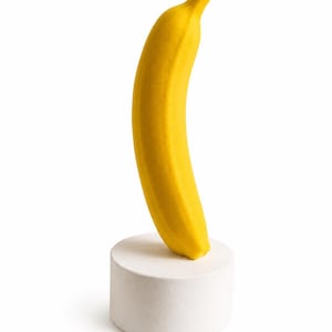 Può includere: Una scultura di banana giallo brillante posizionata verticalmente su una solida base cilindrica bianca. La banana presenta una consistenza liscia e opaca e una forma curva, mentre la base è un semplice cilindro bianco a superficie piatta.