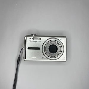 Puede incluir: Cámara digital plateada Olympus X-845 con lente y correa para la muñeca. La cámara tiene un zoom óptico de 5x y 8,0 megapíxeles. La cámara está sobre un fondo blanco.