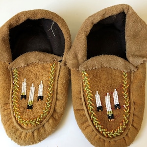 Puede incluir: Un par de mocasines de cuero marrón con diseños de cuentas. Los mocasines tienen un patrón de cuentas blancas, negras y naranjas alrededor de los bordes y un diseño de cuentas blancas, negras y naranjas en la punta.