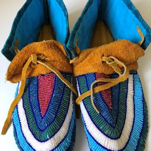 Puede incluir: Un par de mocasines hechos a mano con intrincado trabajo de abalorios. Los mocasines presentan un vibrante patrón de cuentas azules, blancas, verdes y rojas. Tienen la parte superior de cuero marrón y puños azules. Cordones de cuero aseguran la parte superior.