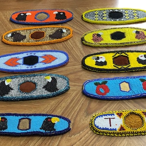 May include: A selection of beaded oval accessories with diverse designs. The beadwork incorporates various colours, patterns, and imagery, including birds, geometric shapes, and text. These accessories are likely for decorative or ceremonial use, with intricate bead detailing.