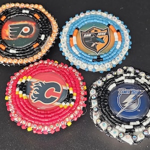 May include: Four round beaded pins featuring hockey team logos. The pins include logos for the Philadelphia Flyers, San Jose Sharks, Calgary Flames, and Tampa Bay Lightning. Each pin is surrounded by colorful beads.