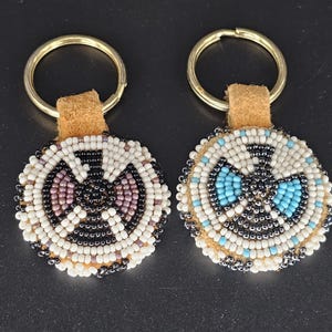 May include: Two beaded keychains with gold-colored key rings. One keychain features a design in white, black, and purple beads. The other has a design in white, black, and turquoise beads. Both have a tan leather backing.